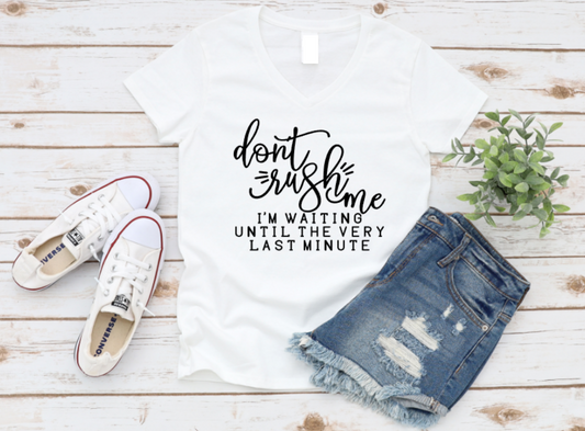 White t-shirt with the quote “Don’t rush me, I’m waiting until the very last minute” in bold white text, styled with Converse shoes and distressed denim shorts on a white wooden background.