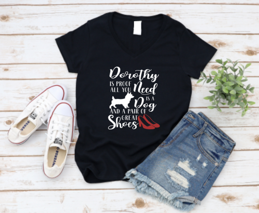 Black t-shirt with bold black and red text that reads "Dorothy is proof all you need is a dog and a pair of great shoes" featuring a dog silhouette and red high heels, styled with denim shorts and sneakers on a wooden background.
