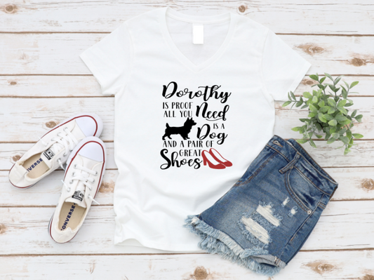 White t-shirt with bold black and red text that reads "Dorothy is proof all you need is a dog and a pair of great shoes" featuring a dog silhouette and red high heels, styled with denim shorts and sneakers on a wooden background.