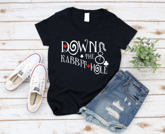 Flat lay of a black v-neck t-shirt that reads “Down the Rabbit Hole” with whimsical fonts and playing card suit symbols, styled with denim shorts, white Converse sneakers, and a plant on a wooden background.