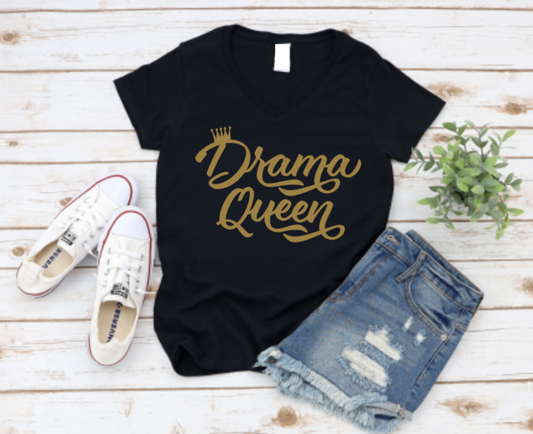 Black t-shirt with “Drama Queen” in bold gold script, styled with ripped denim shorts and white Converse sneakers on a light wood background.