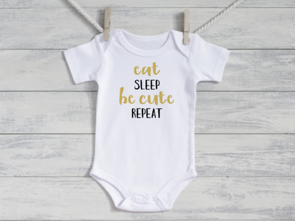 White baby onesie with gold text 'eat sleep be cute repeat' on a wooden background