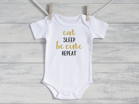 White baby onesie with gold text 'eat sleep be cute repeat' on a wooden background