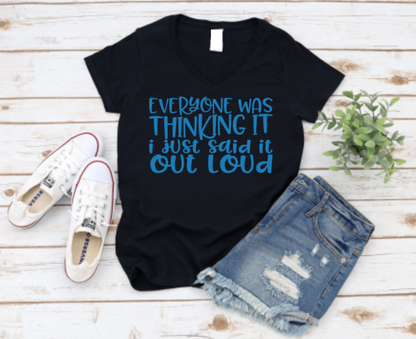 Black v-neck t-shirt with bold blue text that reads “Everyone was thinking it, I just said it out loud,” styled with ripped denim shorts and white Converse sneakers on a wooden background.