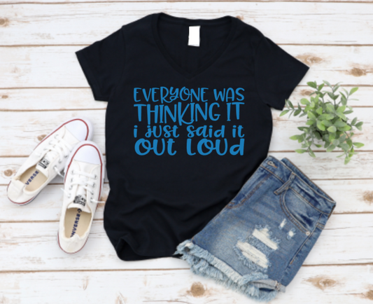 Black v-neck t-shirt with bold blue text that reads “Everyone was thinking it, I just said it out loud,” styled with ripped denim shorts and white Converse sneakers on a wooden background.