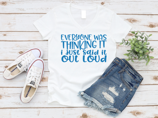 White v-neck t-shirt with bold blue text that reads “Everyone was thinking it, I just said it out loud,” styled with ripped denim shorts and white Converse sneakers on a wooden background.