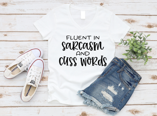 Flat lay of a white V-neck t-shirt with the phrase "Fluent in Sarcasm and Cuss Words" in bold white text, styled with denim shorts and white Converse sneakers on a wooden background.