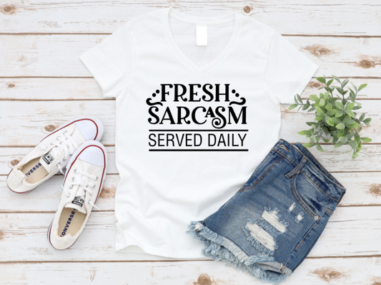 White t-shirt laid flat with black text that reads “Fresh Sarcasm Served Daily,” styled with distressed denim shorts and white Converse sneakers on a light wooden background.