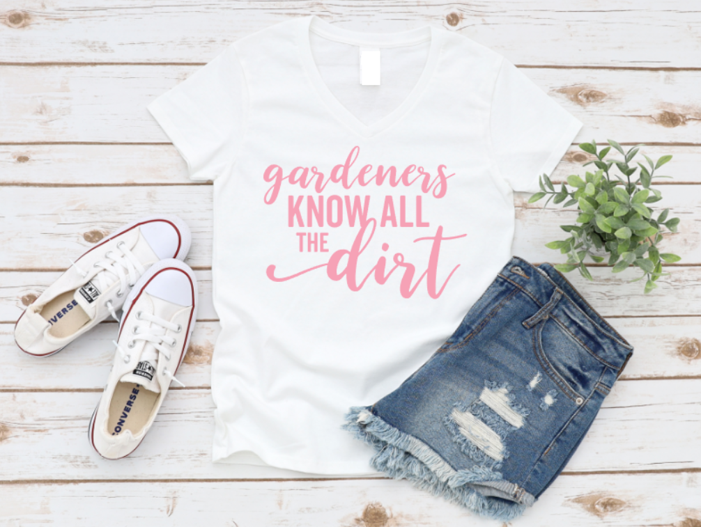 White t-shirt with 'gardeners know all the dirt' text, white sneakers, and denim shorts on a wooden surface.