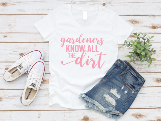 White t-shirt with 'gardeners know all the dirt' text, white sneakers, and denim shorts on a wooden surface.