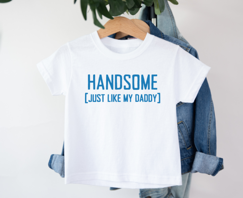 White t-shirt with 'HANDSOME [JUST LIKE MY DADDY]' text on a hanger with denim jacket.