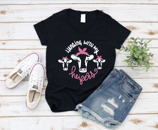 Black t-shirt with cow graphic and text 'Laughing with my heifers' on a wooden surface with shoes and shorts.