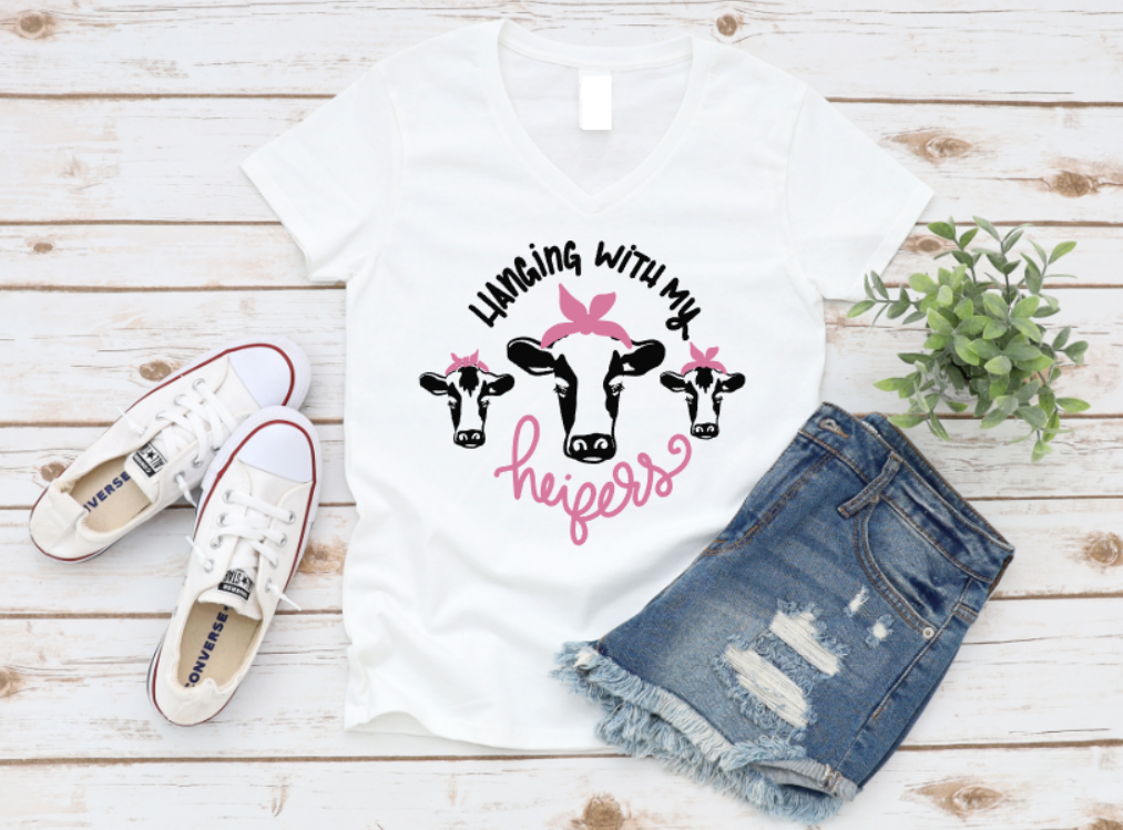 White tank top with 'hanging with my heifers' design, denim shorts, and white sneakers on a wooden surface.