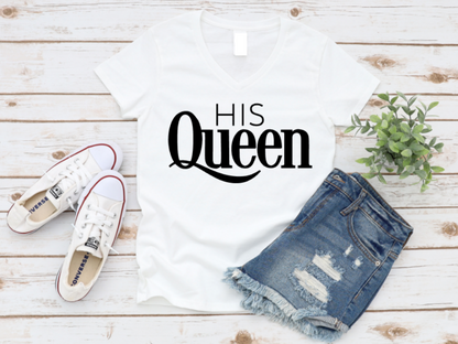 White top with 'His Queen' text, white sneakers, and denim shorts on a wooden surface.