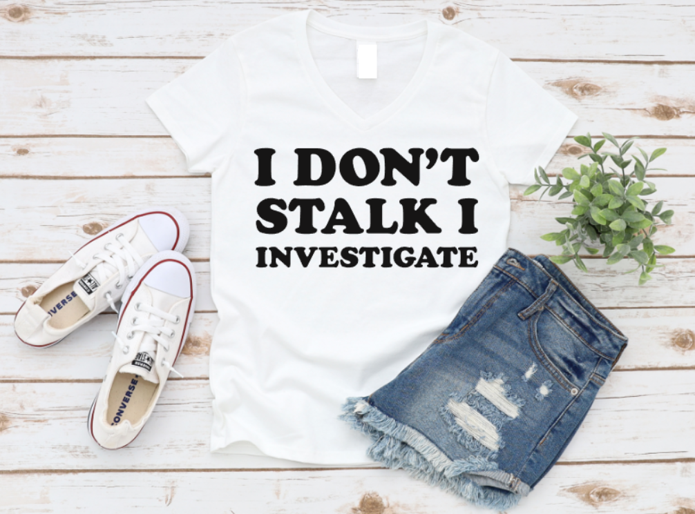 White t-shirt with 'I DON'T STALK I INVESTIGATE' text, white sneakers, and denim shorts on a wooden surface.