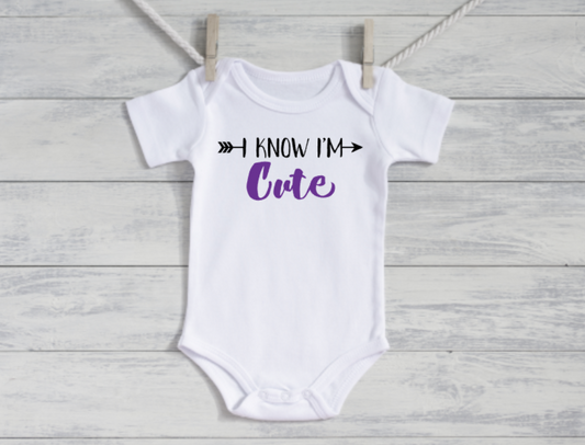 White baby onesie with 'I Know I'm Cute' text on a wooden surface