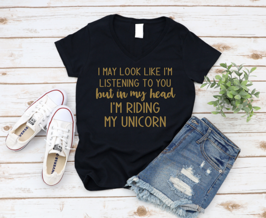 Black t-shirt with gold text 'I may look like I'm listening to you but in my head I'm riding my unicorn' on a wooden surface with jeans and shoes.