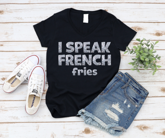 Flat lay of a white v-neck t-shirt with bold black text that reads "I Speak French Fries," styled with denim shorts, white Converse sneakers, and a small green plant on a wooden background.