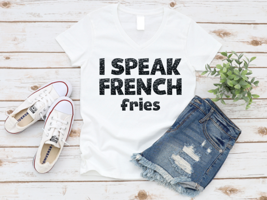 Flat lay of a white v-neck t-shirt with bold black text that reads "I Speak French Fries," styled with denim shorts, white Converse sneakers, and a small green plant on a wooden background.
