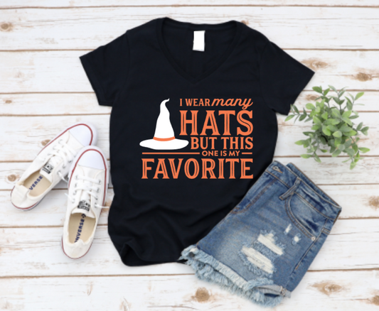 Black t-shirt with 'I wear many hats but this one is my favorite' text and graphics, paired with denim shorts and white sneakers on a wooden surface. Shop at www.missmafia.com