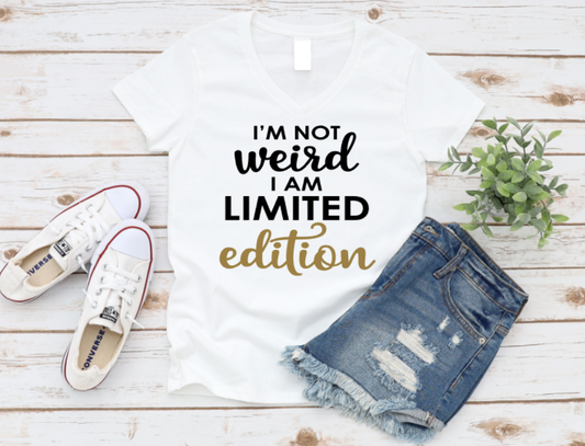 White t-shirt with 'I'm not weird, I am limited edition' text, white sneakers, and denim shorts on a wooden surface. www.missmafia.com