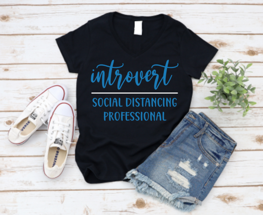 Black t-shirt with 'Introvert Social Distancing Professional' text, paired with white sneakers and denim shorts on a wooden surface. www.missmafia.com