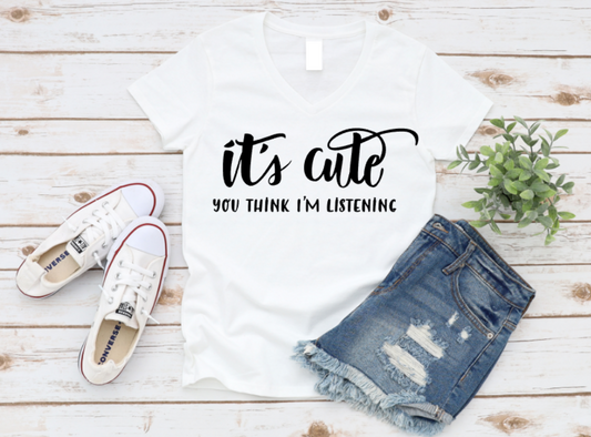 White t-shirt with 'it's cute you think I'm listening' text, white sneakers, and denim shorts on a wooden surface. www.missmafia.com