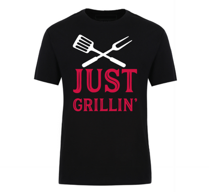 Black t-shirt with bold text “Just Grillin’” featuring a grilling spatula and flame graphic, laid out flat with casual summer essentials—ideal for BBQ lovers and backyard chefs.