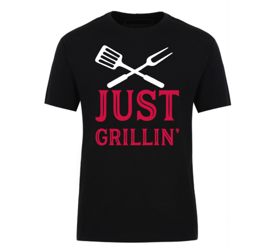Black t-shirt with bold text “Just Grillin’” featuring a grilling spatula and flame graphic, laid out flat with casual summer essentials—ideal for BBQ lovers and backyard chefs.