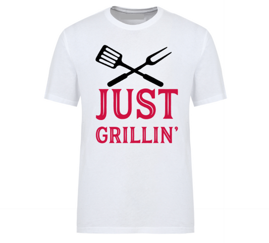 White t-shirt with bold text “Just Grillin’” featuring a grilling spatula and flame graphic, laid out flat with casual summer essentials—ideal for BBQ lovers and backyard chefs.