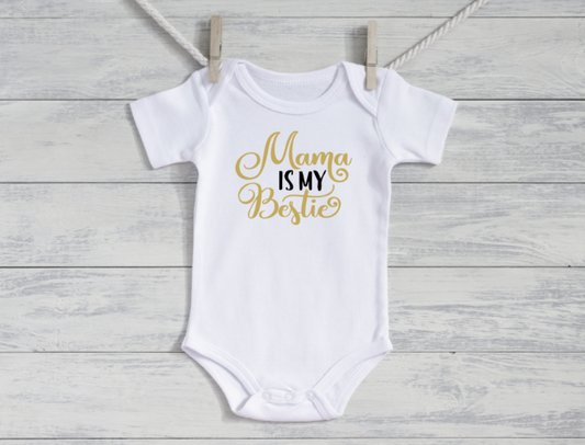 White baby onesie with 'Mama is my Bestie' text in gold on a wooden surface.