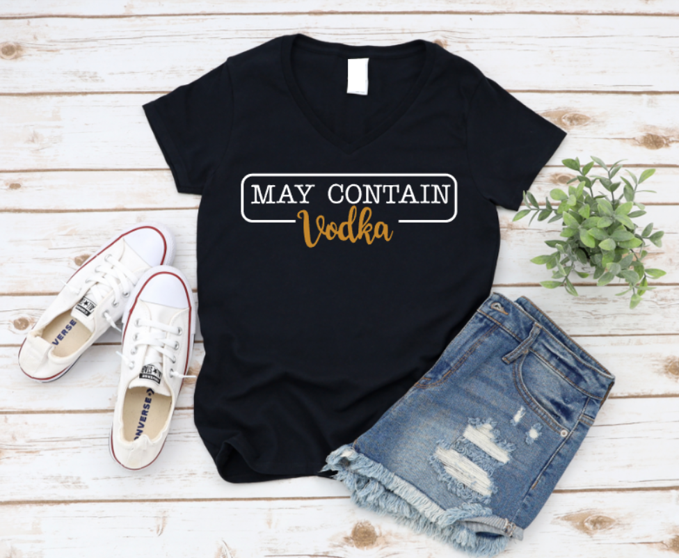 Flat lay of a black t-shirt with the phrase “May Contain Vodka” in black and gold text, styled with denim shorts and white Converse sneakers on a wooden background.