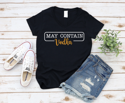 Flat lay of a black t-shirt with the phrase “May Contain Vodka” in black and gold text, styled with denim shorts and white Converse sneakers on a wooden background.