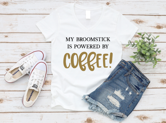 White t-shirt with 'My broomstick is powered by coffee!' text, paired with denim shorts and white sneakers on a wooden surface. www.missmafia.com
