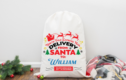 White Santa sack with festive text and graphics on a wooden floor with Christmas decorations.