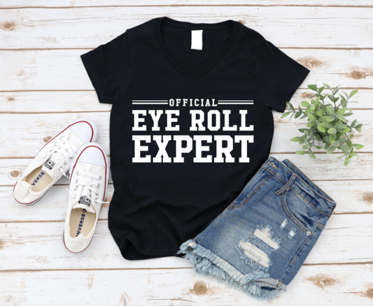 Flat lay of a Black t-shirt with bold black text reading “Official Eye Roll Expert,” styled with denim shorts, Converse sneakers, and a plant on a rustic wooden background.