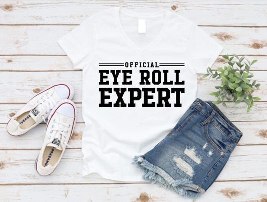 Flat lay of a white t-shirt with bold black text reading “Official Eye Roll Expert,” styled with denim shorts, Converse sneakers, and a plant on a rustic wooden background.