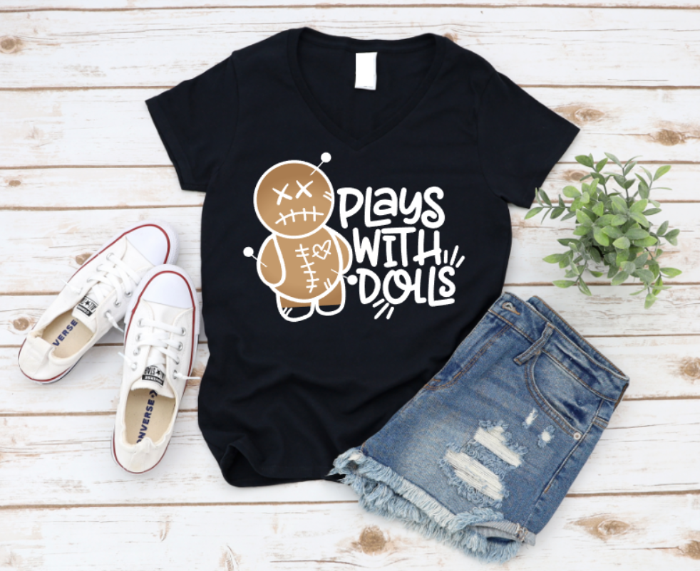 Black t-shirt with a playful voodoo doll graphic and bold white text that reads 'Plays With Dolls'. Styled flat lay with denim shorts, white Converse sneakers, and greenery on a light wooden background. www.missmafia.com