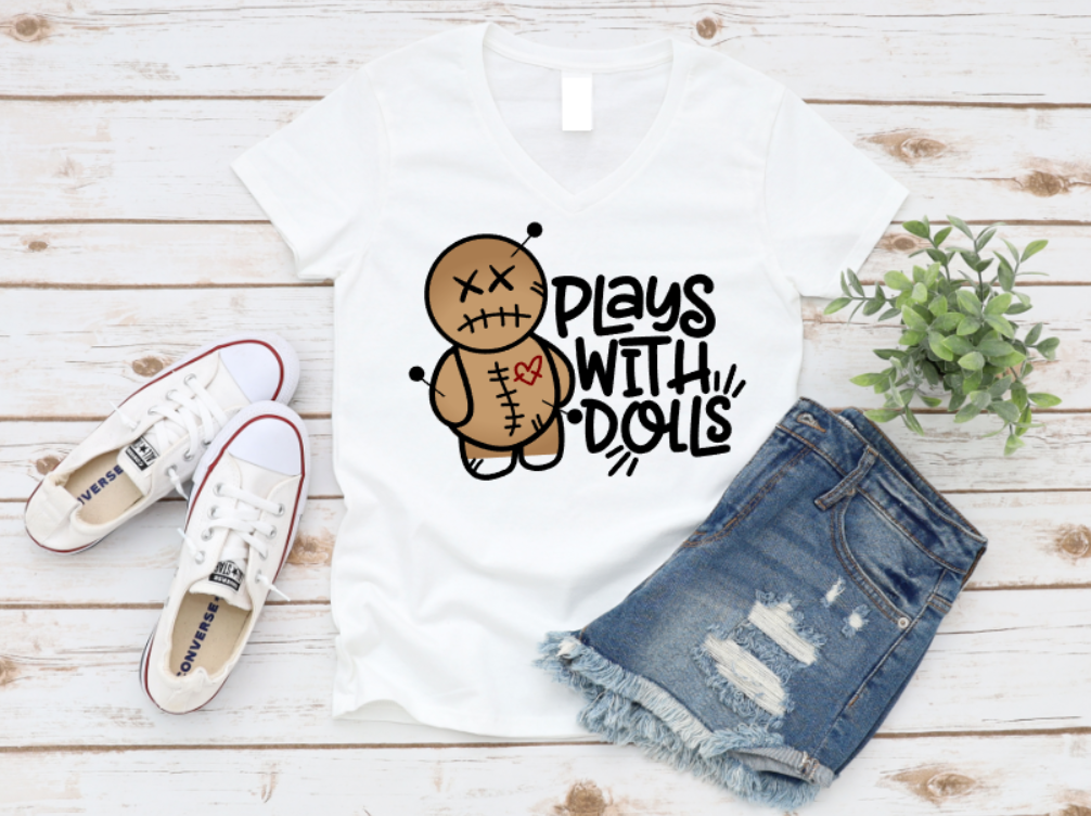 White t-shirt with a playful voodoo doll graphic and bold white text that reads 'Plays With Dolls'. Styled flat lay with denim shorts, white Converse sneakers, and greenery on a light wooden background. www.missmafia.com