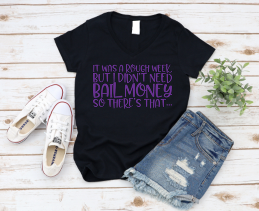 Black t-shirt with purple "It was a rough week but I didn’t need bail money, so there’s that..." text, styled with denim shorts and white sneakers on a wooden background.