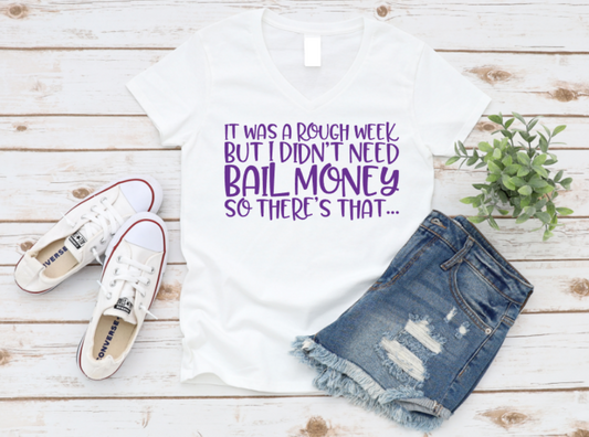 White t-shirt with purple "It was a rough week but I didn’t need bail money, so there’s that..." text, styled with denim shorts and white sneakers on a wooden background.