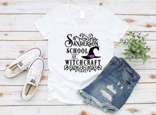 White t-shirt with white gothic-style text that reads "Sanderson School of Witchcraft – 1692" and features a purple and green witch hat illustration. Shirt is styled flat lay with denim shorts, white Converse sneakers, and a small green plant on a light wood background. www.missmafia.com