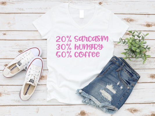 Flat lay of a white t-shirt with pink text reading "20% sarcasm, 30% hungry, 50% coffee" displayed on a white wooden background with white sneakers, denim shorts, and a green plant.