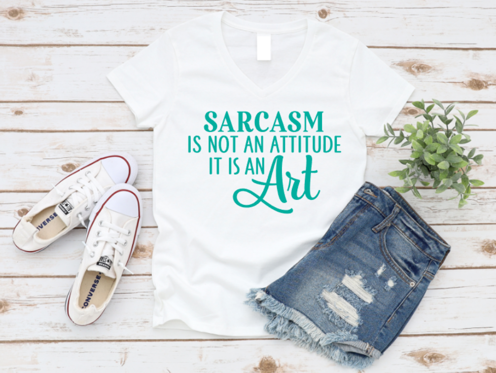 White t-shirt with 'Sarcasm is not an attitude, it is an art' text, paired with denim shorts and white sneakers on a wooden surface. www.missmafia.com