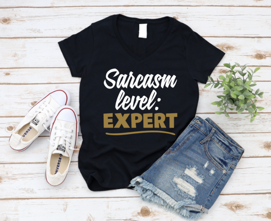 Black t-shirt with 'Sarcasm level: Expert' text, white sneakers, and denim shorts on a wooden surface. www.missmafia.com