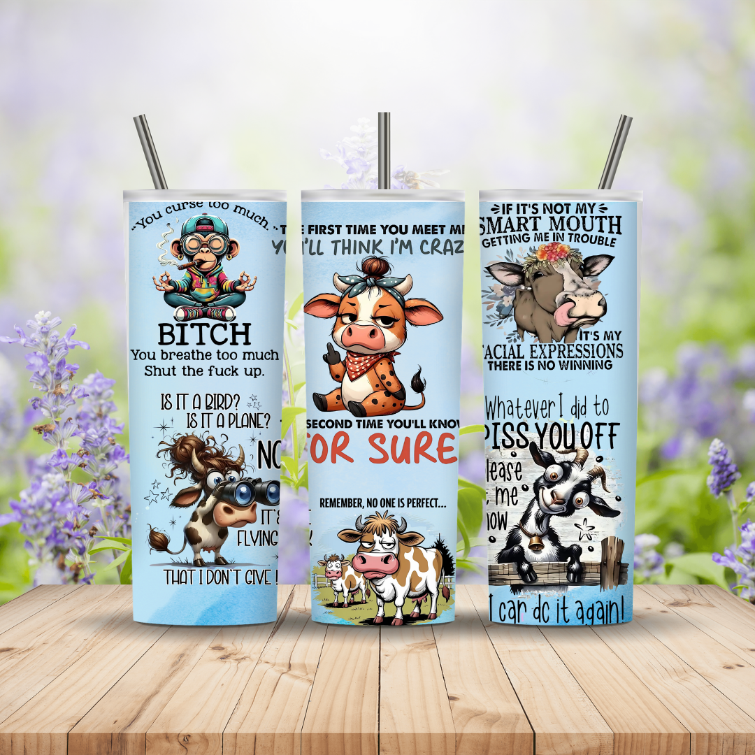 Three tumblers with cow designs and humorous text on a wooden surface with a blurred floral background.