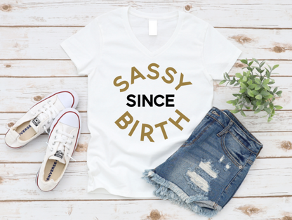 Flat lay of a white t-shirt with the phrase “Sassy Since Birth” printed in bold gold and white text, styled with distressed denim shorts, white Converse sneakers, and a small green plant on a white wooden background. | www.missmafia.com