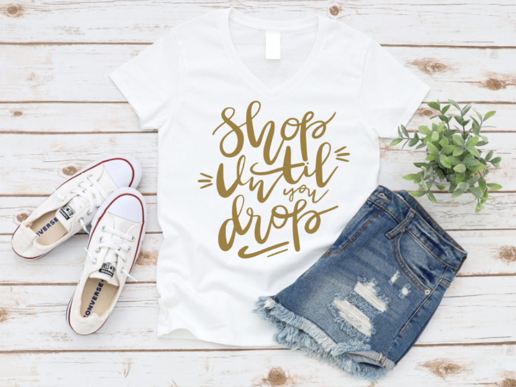 White tote bag with 'Shop till you drop' text, white sneakers, denim shorts, and a plant on a wooden surface. WWW.MISSMAFIA.COM