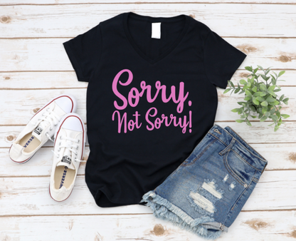 Flat lay of a black t-shirt with pink text that reads “Sorry. Not Sorry!”, styled with denim shorts, white Converse sneakers, and a small green plant on a white wooden background. www.missmafia.com