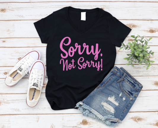 Flat lay of a black t-shirt with pink text that reads “Sorry. Not Sorry!”, styled with denim shorts, white Converse sneakers, and a small green plant on a white wooden background. www.missmafia.com